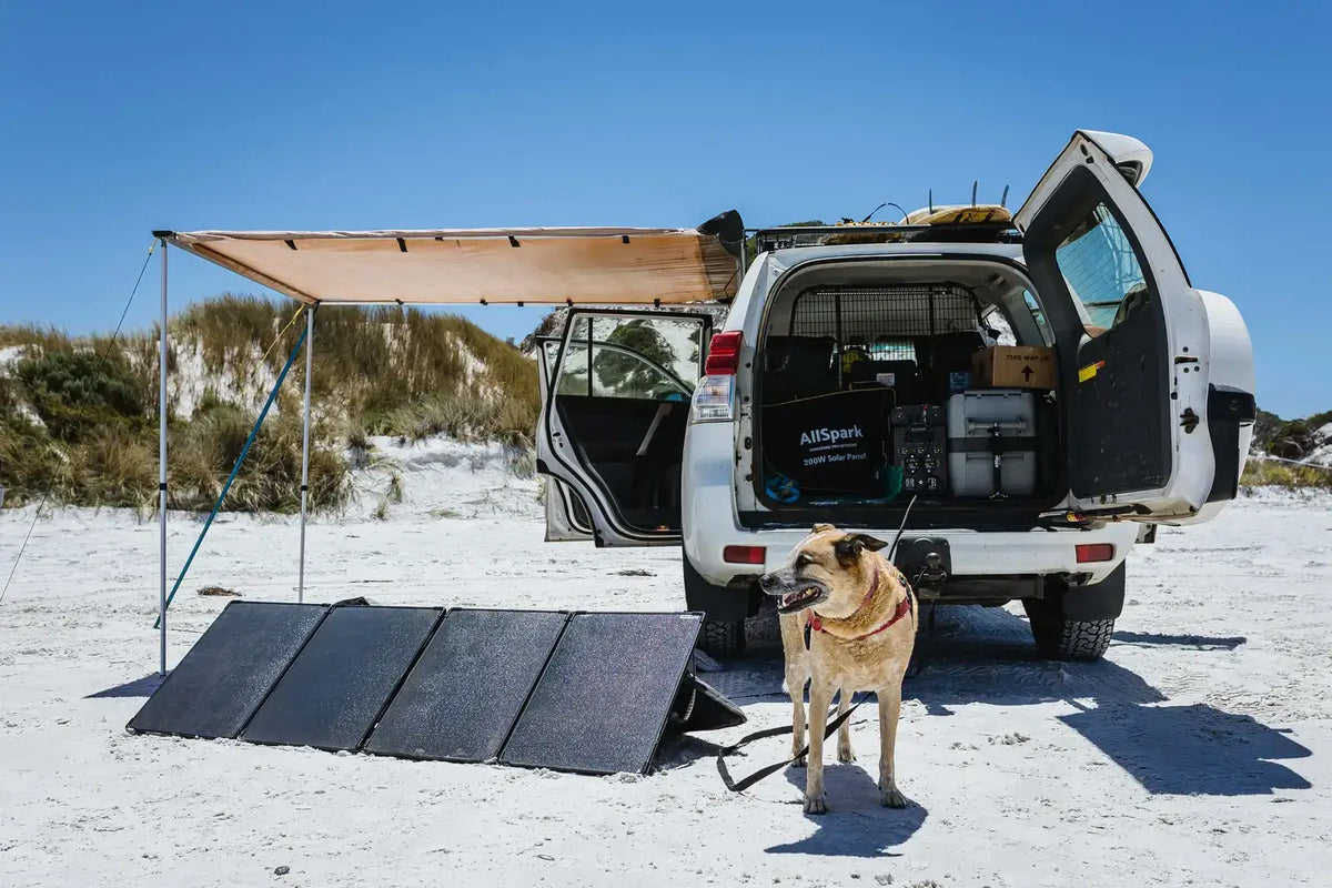 4WD set up on a beach with an awning, folding solar panels, an AllSpark battery in the boot and a dog on a lead